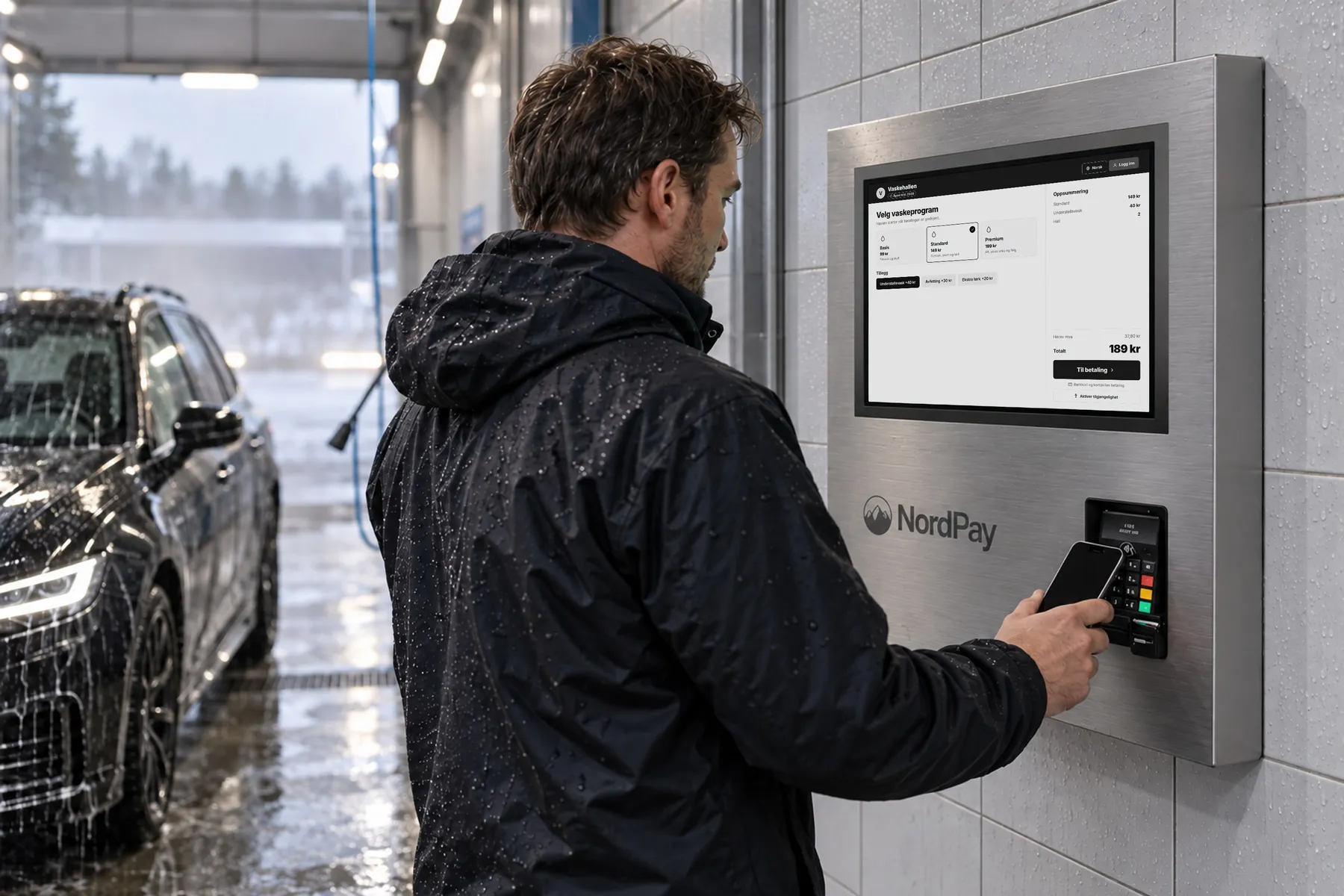 Mann betaler med mobilen på en veggmontert betalingsautomat i en vaskehall